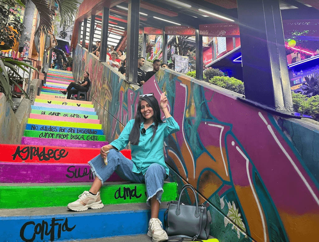 Roxeus sitting on colorful stairs at Comuna 13 in Medellín, Colombia, with vibrant street art in the background