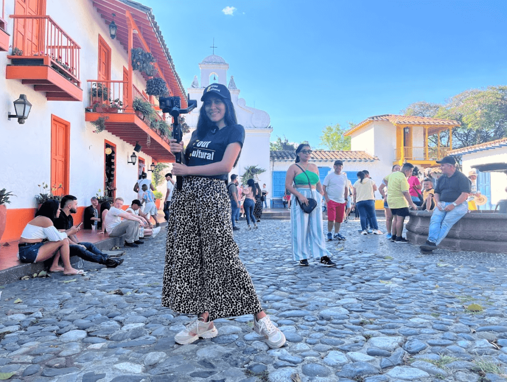 Roxeus at Pueblito Paisa in Medellín, Colombia, holding a camera with tourists in the background