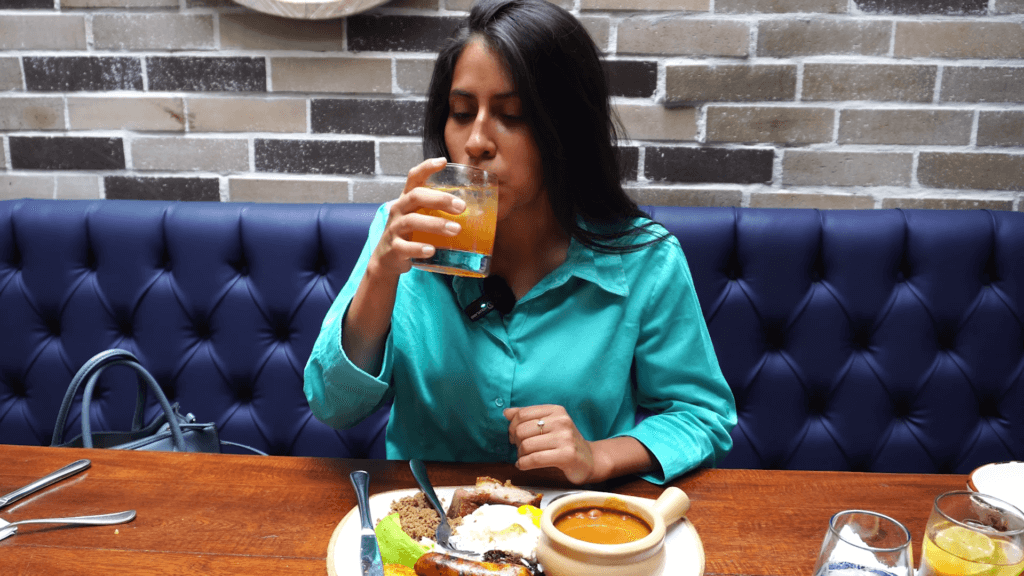 Woman drinking a glass of refajo and a bandeja paisa while sitting in a restaurant.